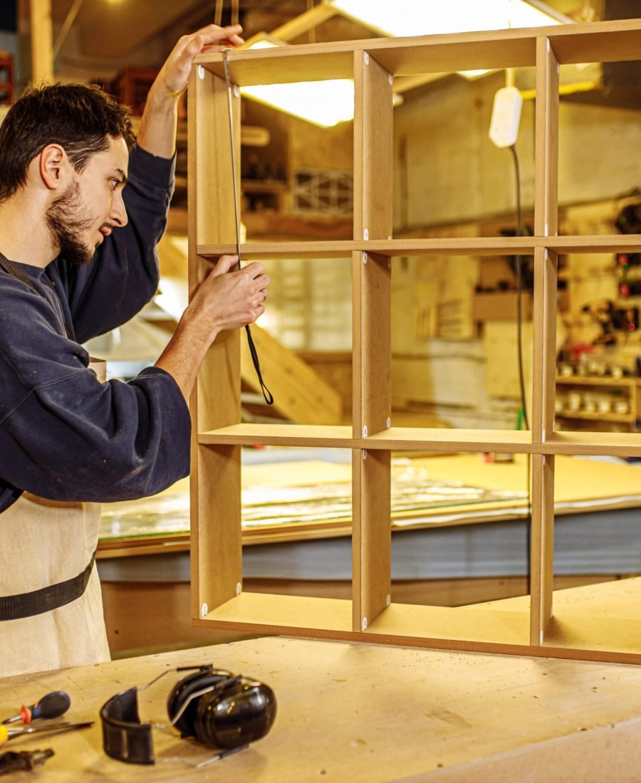 custom-made furniture concept. handicraft carpentry. young caucasian woodworker measuring a hand made wooden shelves using tape-line in factory